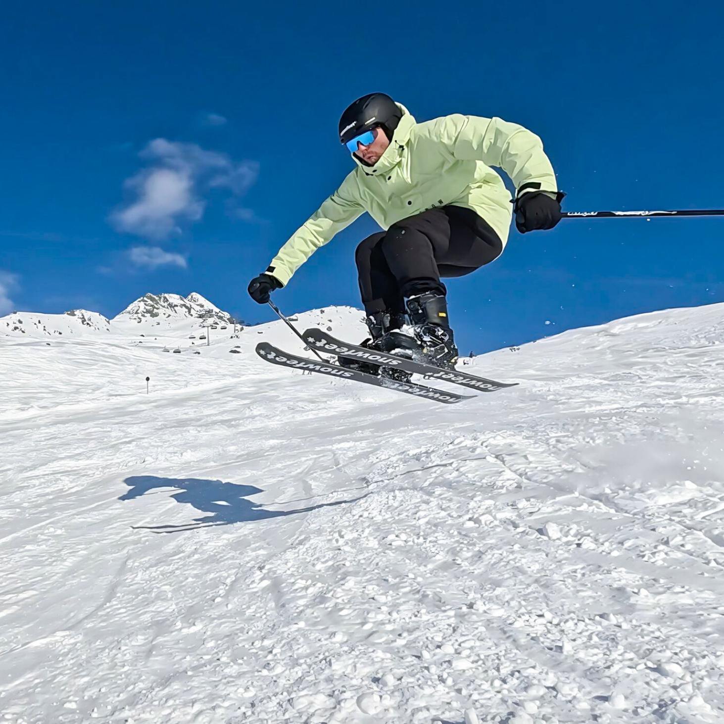 Skier in a yellow jacket and black helmet skiing down a snowy slope with a clear blue sky.