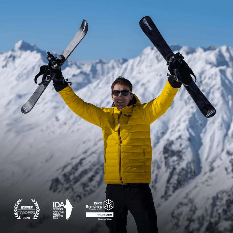 Person in a yellow jacket holding skis and snowboard against a snowy mountain backdrop