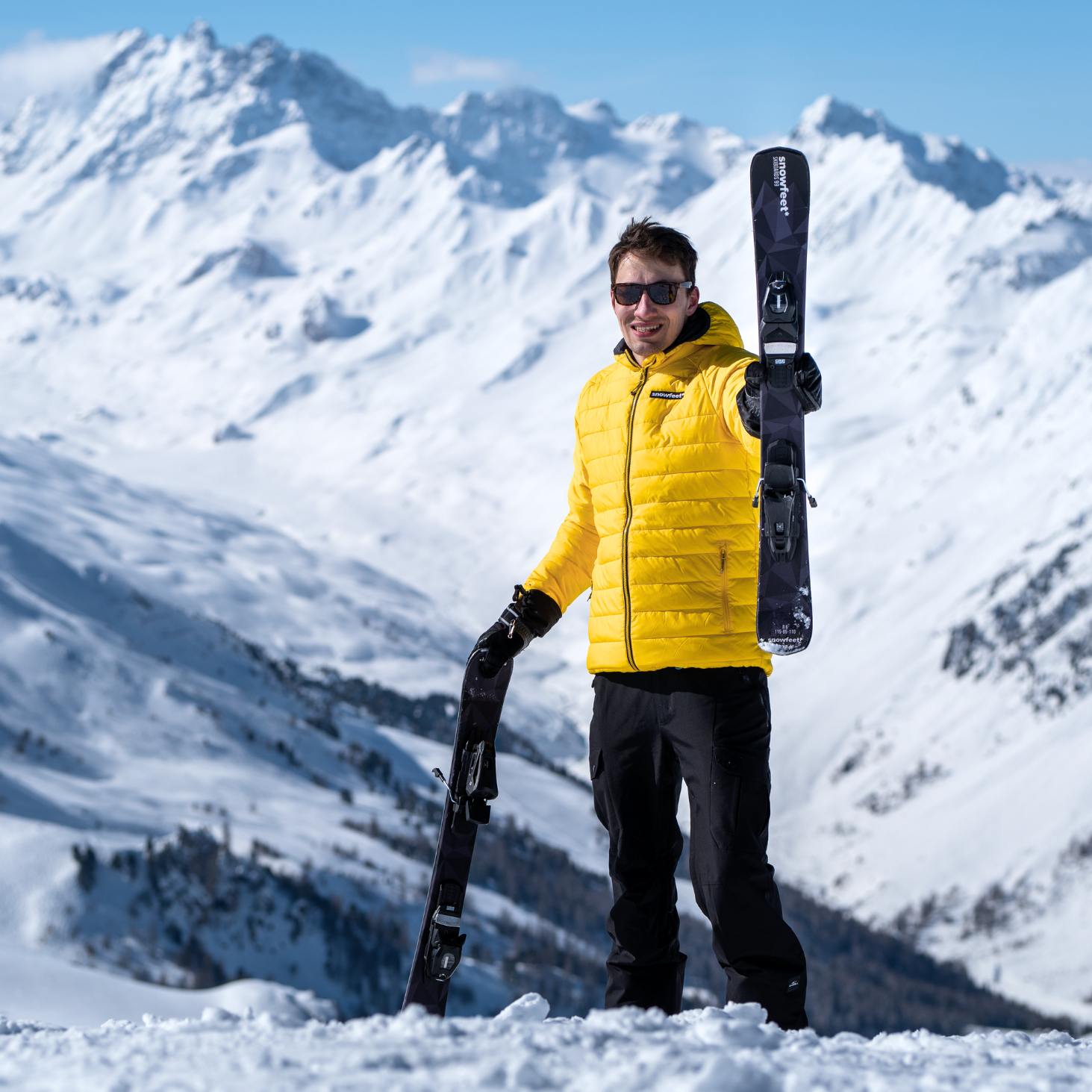 Person in a yellow jacket holding skis against a snowy mountain backdrop. skiblades* Short Skis | 99 CM | Retro | Snowboard Bindings |  Skiblades Skiboards Snowblades - snowfeet*